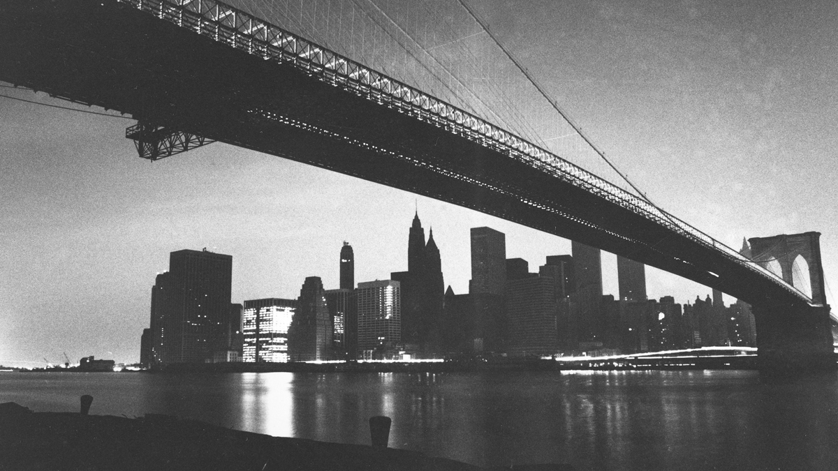 The lights of lower Manhattan are dark as a result of an electrical power failure in this view from under the Brooklyn Bridge on Wednesday, July 13, 1977. One building at left is lit with emergency power and a stream of light comes from the headlights of moving vehicles on the FDR Drive and on the bridge. (AP Photo/Ray Stubblebine)