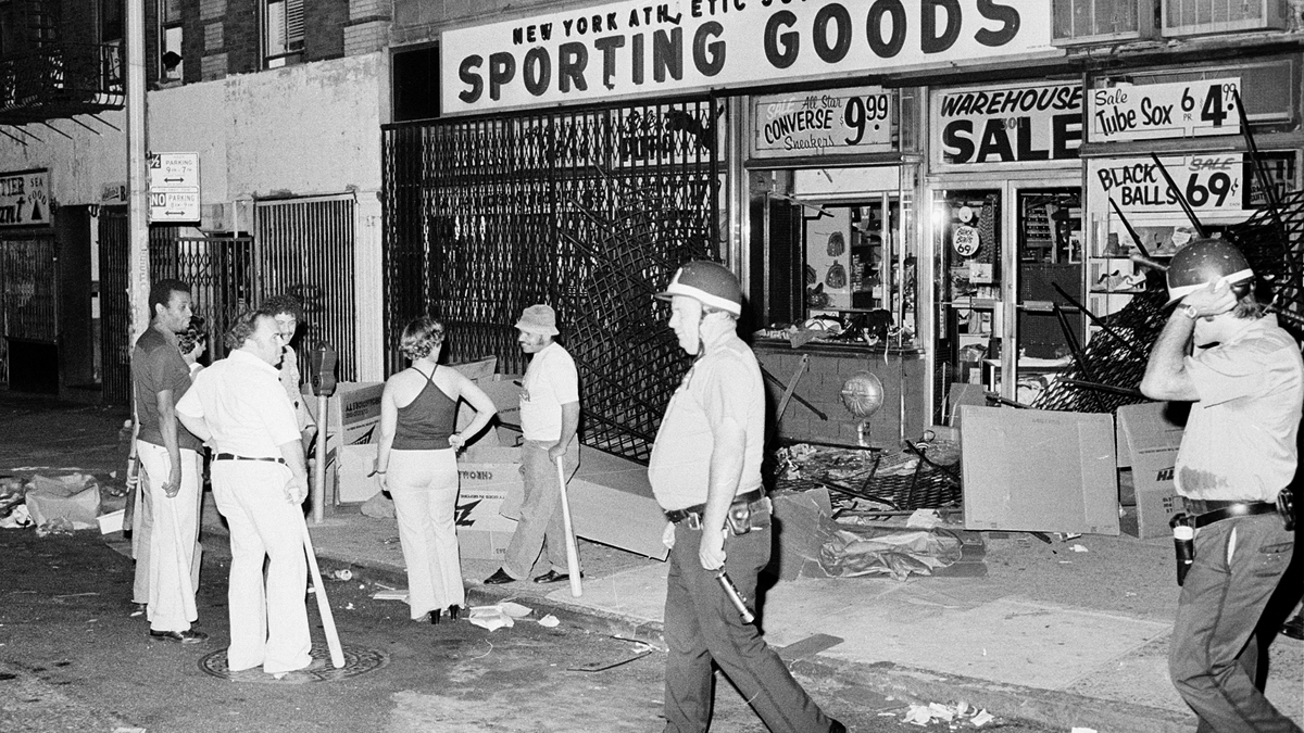 Owners and employees of a sporting goods store stand guard outside with baseball bats after the store on New York City's Upper West Side was looted during massive power failure, Wednesday night, July 13, 1977. Police patroling the area walk by the store. (AP Photo)