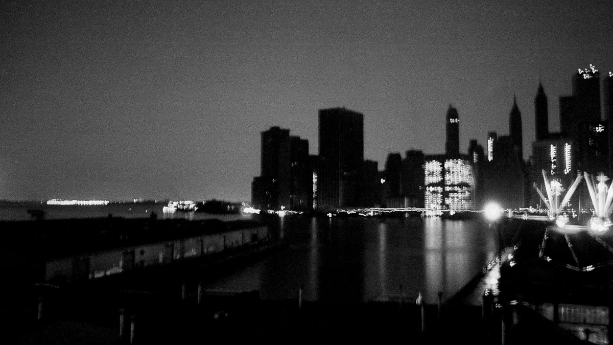 The darkened Manhattan skyline is seen from Brooklyn after the city was hit by a power failure, July 13, 1977. Bright spot at left is the Statue of Liberty; at right is a ship in harbor and a few building lights are on in places with their own auxiliary power. (AP Photo/PB)