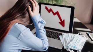 Top view of stressed young woman holding her head in her hands as her laptop screen shows a downward-trending line graph and a stack of surgical masks can be seen next to the laptop.