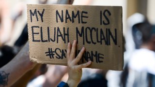 AURORA, CO - JUNE 27: A woman holds up a sign as people rally outside the Aurora Police Department Headquarters to demand justice for Elijah McClain on June 27, 2020 in Aurora, Colorado. On August 24, 2019 McClain was walking home when he was forcibly detained by three Aurora police officers and was injected with ketamine after officers requested assistance from the Aurora Fire Rescue. McClain suffered a heart attack on the way to the hospital that night and died six days later.