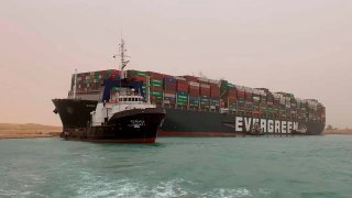 In this photo released by the Suez Canal Authority, a boat navigates in front of a massive cargo ship, named the Ever Green, rear, sits grounded Wednesday, March 24, 2021, after it turned sideways in Egypt’s Suez Canal, blocking traffic in a crucial East-West waterway for global shipping. An investigation into the incident is underway after the ship was freed on March 29, 2021.