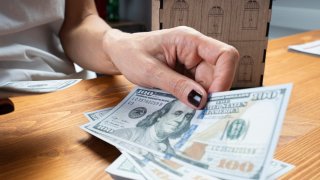 A hand holds $100 bills as a miniature house sits behind it.