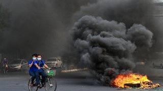 This picture taken on April 3, 2021 shows people cycling past a burning makeshift barricade, erected by protesters demonstrating against the military coup, in Yangon's Tamwe township, Myanmar.