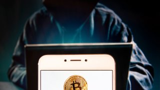 In this photo illustration, a bitcoin logo is seen displayed on an Android smartphone with a hacker in the background.