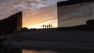 pair of migrant families from Brazil passes through a gap in the border wall