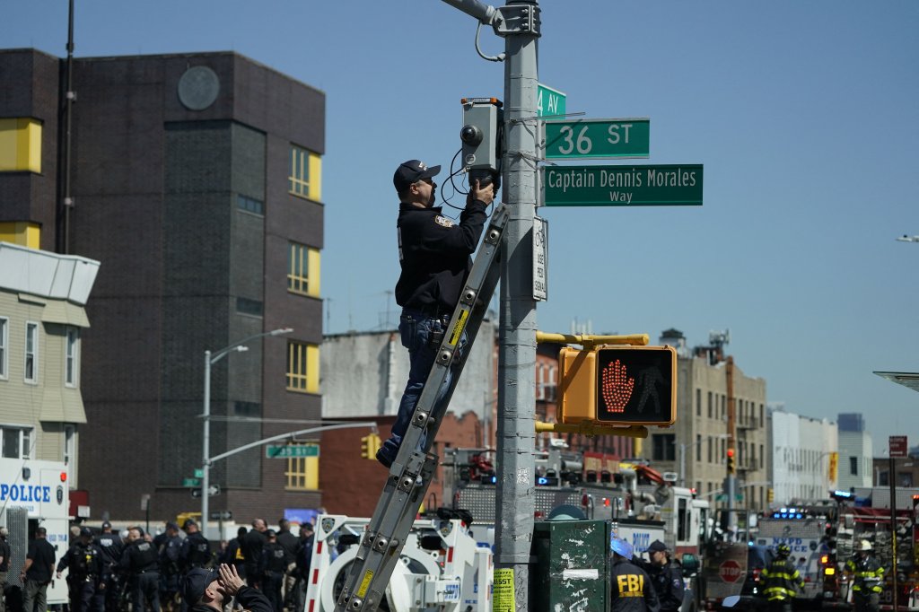 A member of the NYPD retrieves a security camera near a subway station in New York City on April 12, 2022, after at least 16 people were injured during a rush-hour shooting in the Brooklyn neighborhood of Sunset Park.
