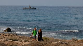 People walking along the beach as a navy vessel patrols the Mediterranean waters off Rosh Hanikra, known in Lebanon as Ras al-Naqura, on the Israeli side of the border between the two countries, on October 7, 2022. Israel and Lebanon have brokered a historic agreement to resolve a long-running maritime border dispute on Tuesday, following months of negotiations mediated by the United States.