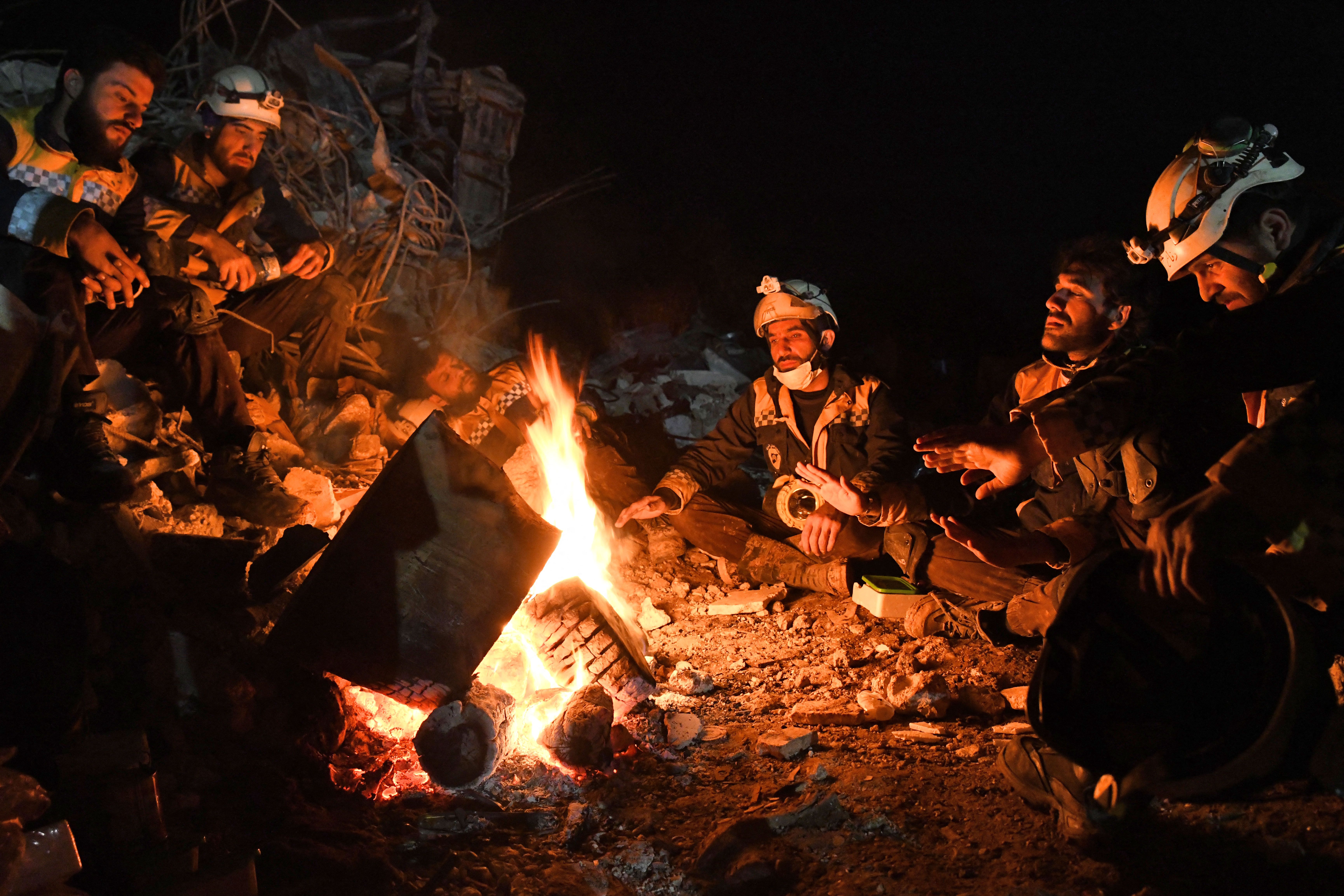 Members of the Syrian civil defense, known as the White Helmets, warm themselves by a fire next to the rubble of a collapsed building in Jinderis, Feb. 7, 2023, as search and rescue operations continue following a deadly earthquake.