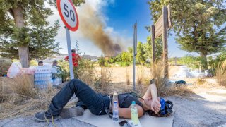 25 July 2023, Greece, Gennadi: A relief worker, who has been on duty for days, has laid down on a piece of cardboard by the side of the road to rest, in the background the plume of smoke from a newly erupted forest fire in the village of Gennadi. Mediterranean heat – thousands of people flee forest fires on Rhodes. In Greece, forest fires are raging in numerous regions. Popular vacation resorts such as the islands of Rhodes and Corfu are also affected. Photo: Christoph Reichwein/dpa