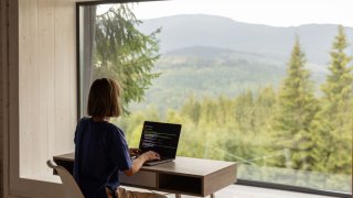 Woman works on laptop in front of panoramic window with mountain view.