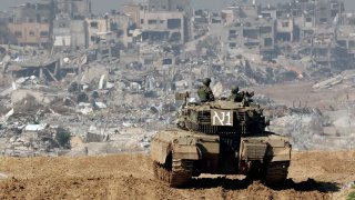 A picture taken from a position in southern Israel along the border with the Gaza Strip on January 19, 2024, shows an Israeli tank rolling along the fence as damaged buildings are see in the Gaza strip amid continuing battles between Israel and the militant group Hamas.