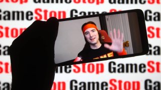 Keith Gill, known on Reddit under the pseudonym DeepF——-Value and as Roaring Kitty, is seen on a fragment of a youtube video displayed on a smartphone screen in front of GameStop logo.