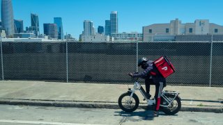 A DoorDash driver on an electric bicycle wearing a cooler backpack with the skyline of San Francisco in the background.