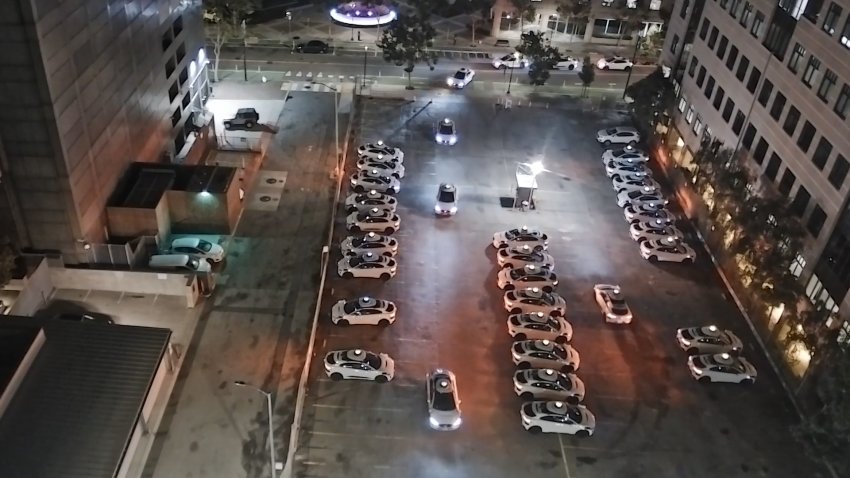 A parking lot at night time filled with more than 30 parked Waymo driverless vehicles and more vehicles entering the lot