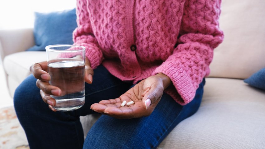 File. Close-up of unrecognizable black woman sitting on couch holding medication/supplements and glass of water