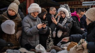 A woman, right, looks at herself on her phone as she and others buy warm winter hats at a vendors shop in the Panjiayuan Market on December 6, 2024 in Beijing, China.
