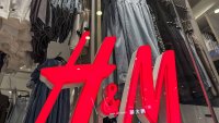 A close-up view of the illuminated red H&M logo in front of a display of women’s summer dresses and casual apparel at a retail store.