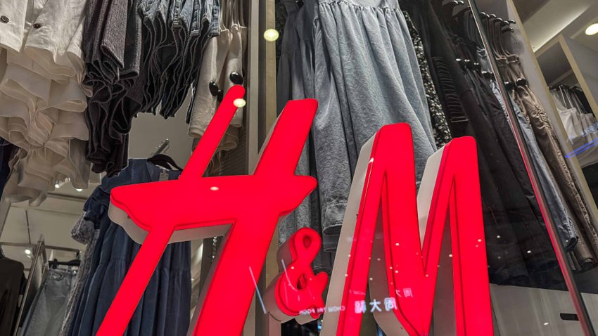 A close-up view of the illuminated red H&M logo in front of a display of women’s summer dresses and casual apparel at a retail store.